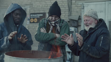 homeless men warming hands above fire on cold day in urban alley