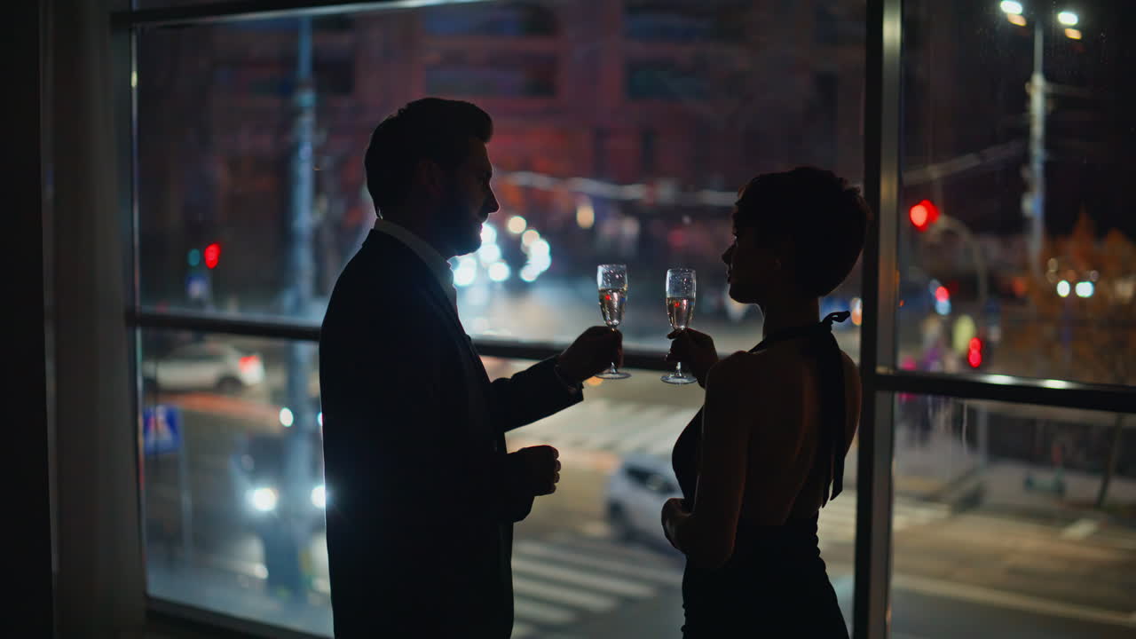 Coppia in appuntamento serale in un hotel di lusso panoramico, visti di spalle. L'uomo porta lo champagne.
