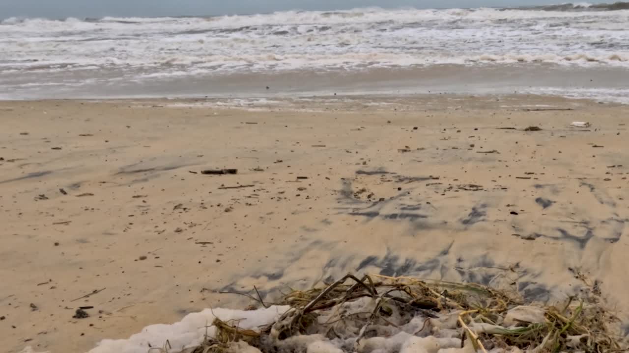 A close-up view of a sandy beach with scattered debris and gentle ocean waves under a cloudy sky.