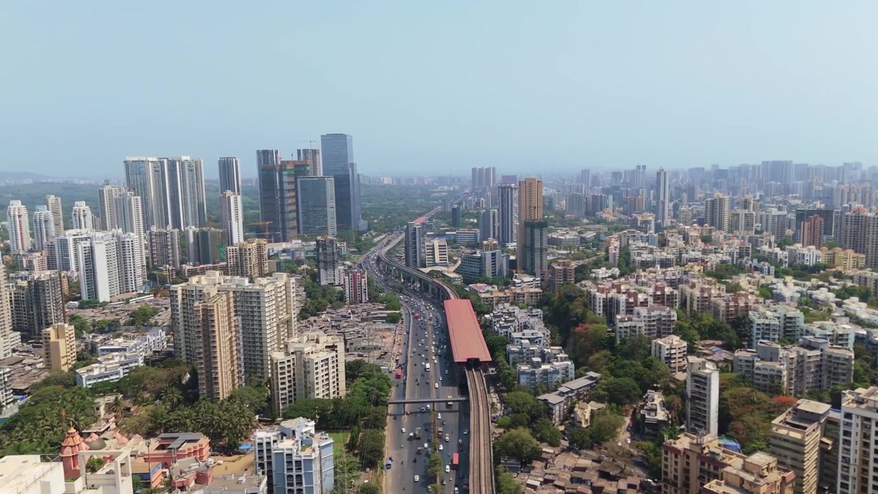 Vasta paisagem urbana de Mumbai, Índia, exibindo arranha-céus residenciais, uma via expressa movimentada com tráfego intenso e uma linha e estação de metrô elevadas em um dia claro e ensolarado.