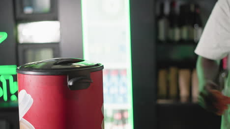 bartender cleaning red trash bin with orange cloth behind bar counter. green neon lighting highlights the background, and shelves are stocked with bottles, creating a festive pub atmosphere