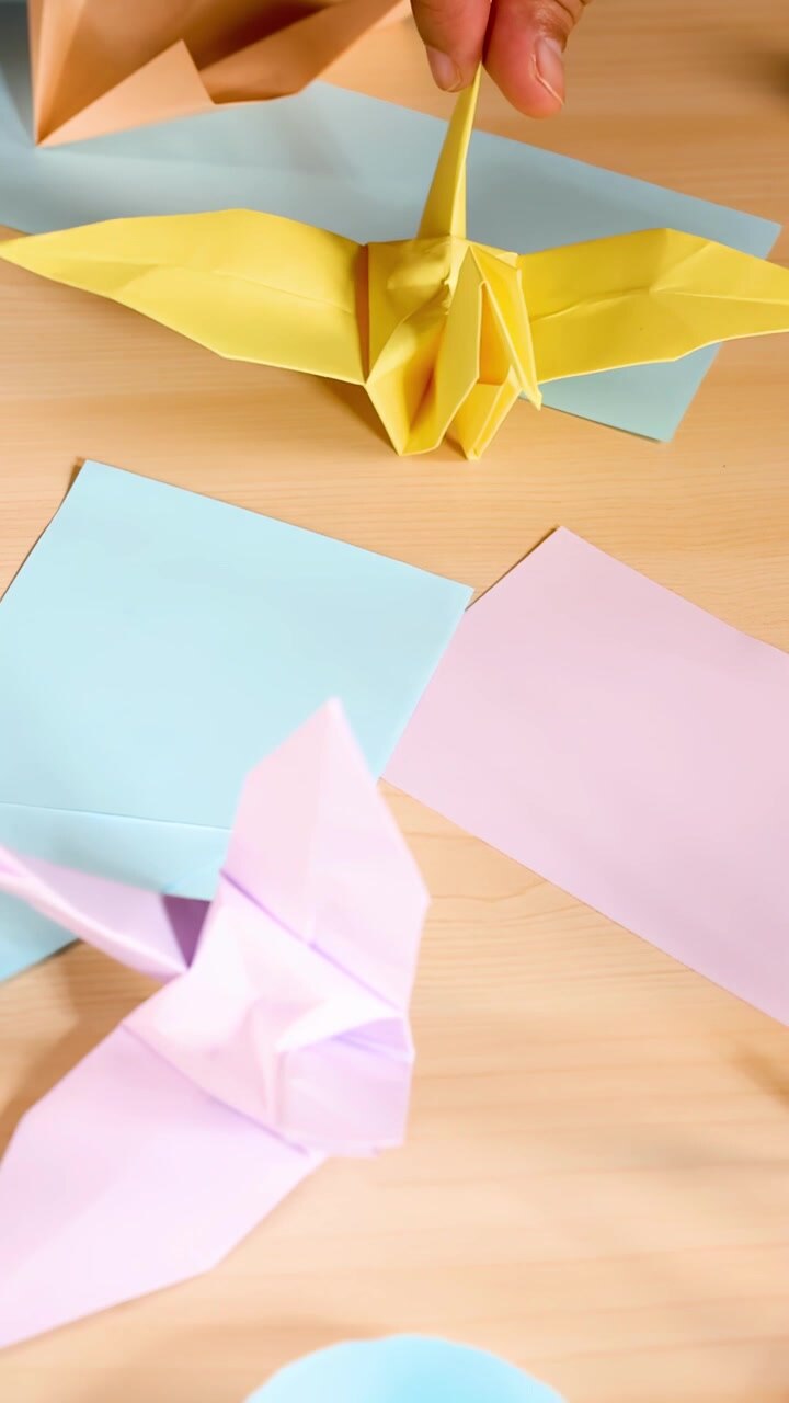 Person folds pastel origami paper crane on wooden desk, bright lighting, close-up perspective