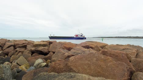 large blue cargo ship leaving port of liepaja , overcast spring day, wide revealing shot from tilt up from stone pier rocks