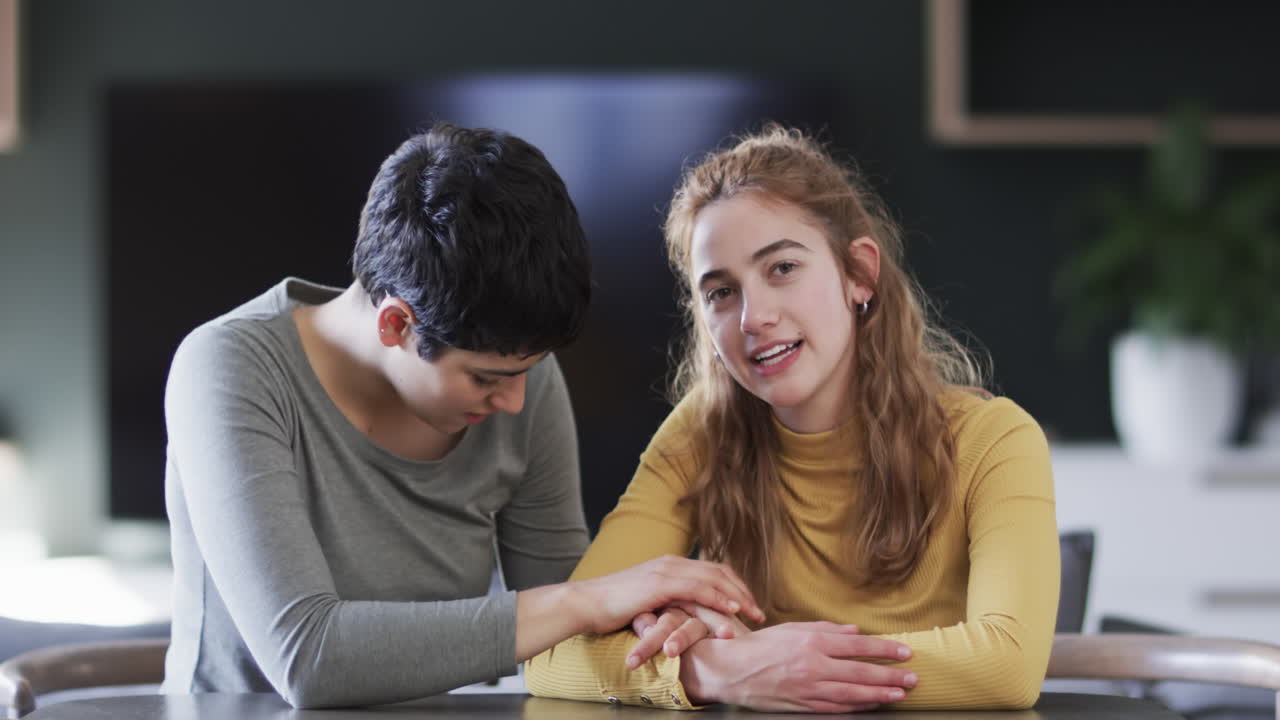 Casal lésbico caucasiano feliz sentado, de mãos dadas e conversando em sala de estar ensolarada.