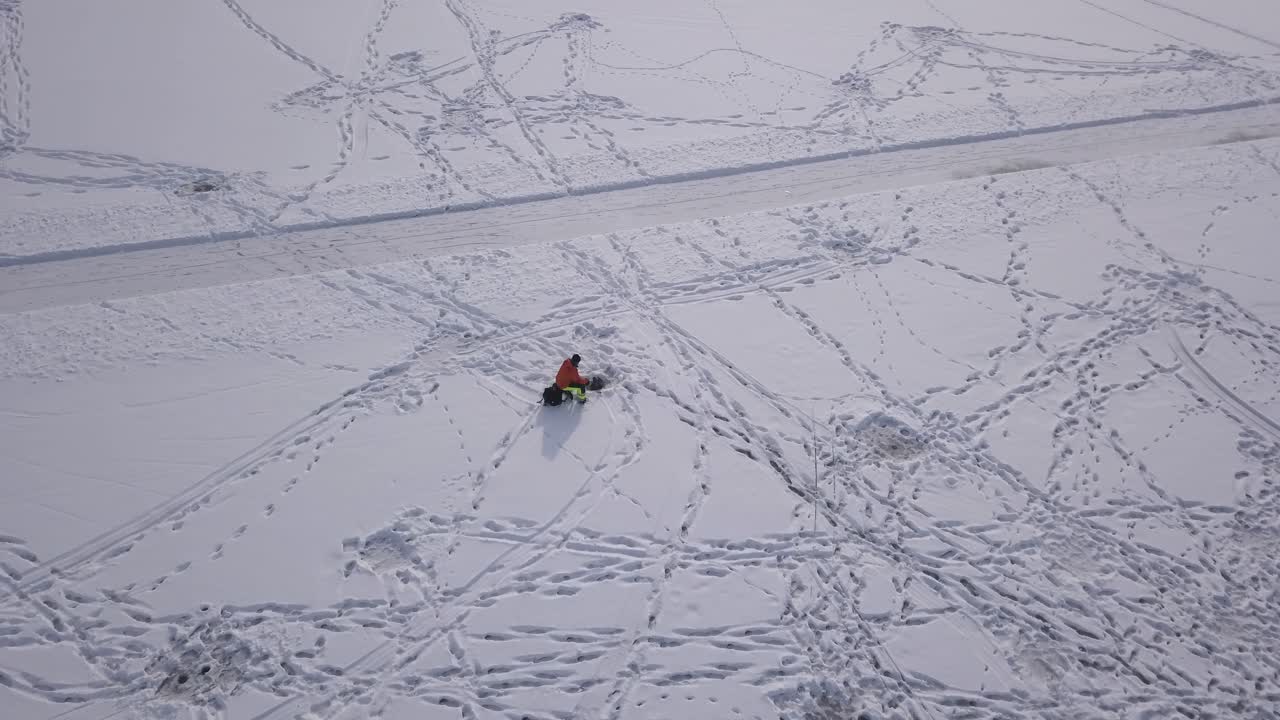 Extreme slow panning aerial view of outdoorsman sitting next to freshly drilled hole while participating in ice fishing.