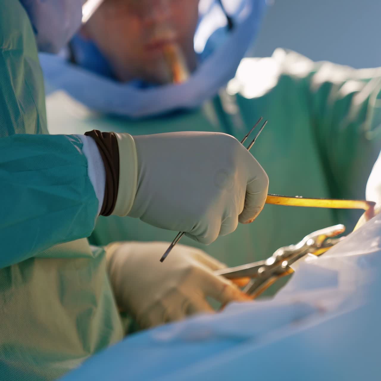 Doctor's hands in latex gloves hold metal tools. Another doctor at backdrop uses metal forceps. Blurred background