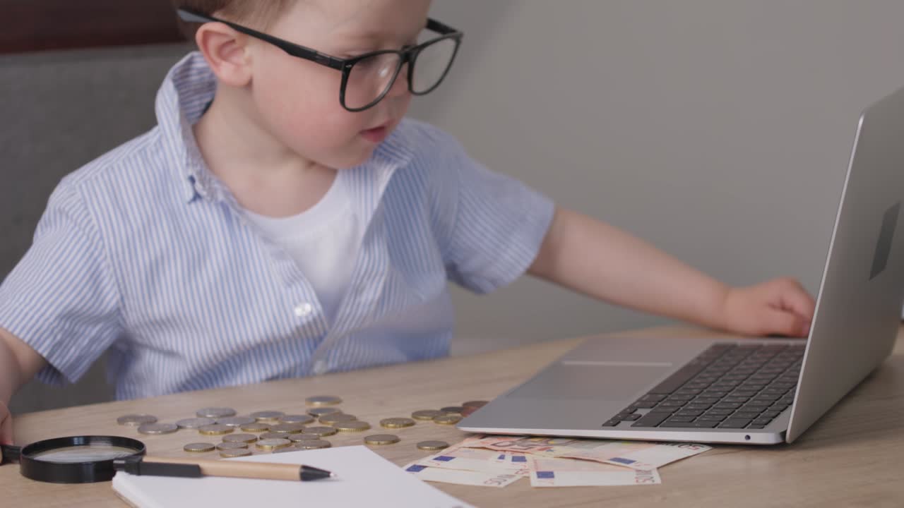 niño niño en camisa azul mecanografiando portátil, dinero billetes de euro, monedas en la mesa
