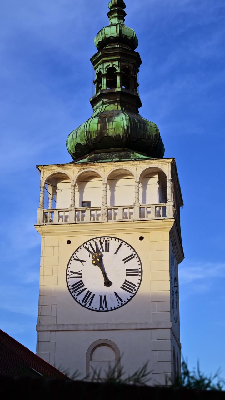 Pystysuuntainen lähikuva valkoisesta kellotornista, jossa on kaarikäytävä ja vihreä sipulikupoli, Mikulov, Tšekki.