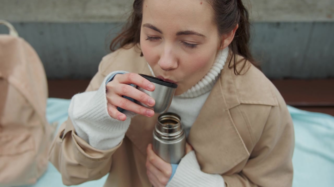 Primo piano di una donna che tiene e beve da un thermos di metallo mentre è seduta su un tessuto azzurro con una borsa beige nelle vicinanze. La donna indossa un cappotto color cammello sopra un dolcevita bianco, con l'attenzione focalizzata unicamente su di lei.