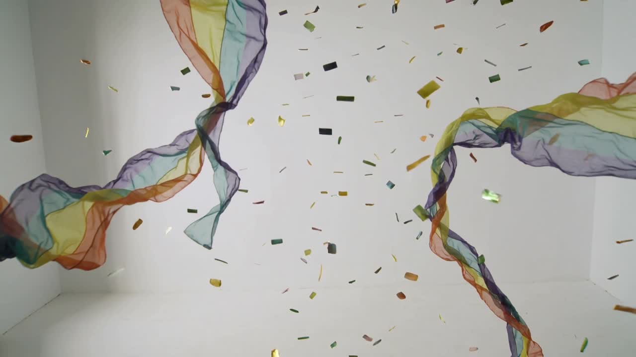En kaskad av två skira regnbågssjalar och glittrande konfetti snurrar och faller ner i en vit kub.