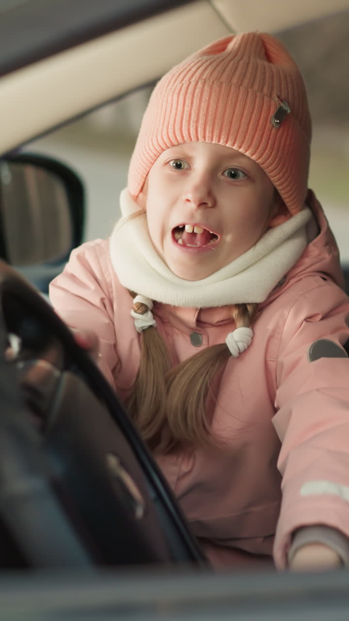 una joven, vestida cálidamente con una gorra rosada y una chaqueta, se sienta en el asiento delantero del pasajero de un coche, girando ansiosamente el volante mientras su padre, sentado a su lado, ríe alegremente