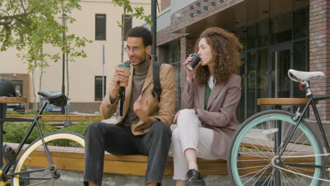 joven estadounidense y mujer con ropa formal sosteniendo café para llevar y hablando mientras se sientan al lado de sus bicicletas en un banco de madera en la ciudad