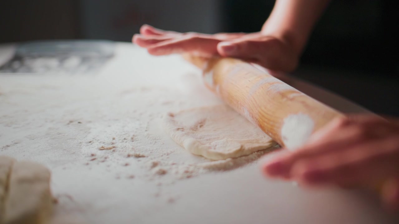 Hands roll dough on floured surface. Person focuses intently on task. Dough stretches under rolling pin. Scene captures home cooking process. Great for recipes, baking tutorials, or food blogs