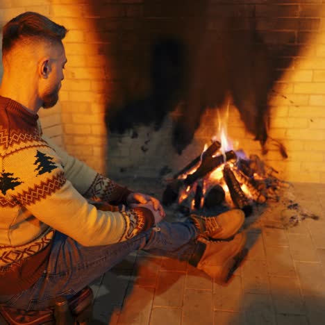 young man warming his hands near the fireplace. man in christmas sweater and jeans sitting by the fire inside the house.