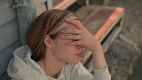 una joven con capucha se sienta con el rostro cubierto por la mano, profundamente en pensamiento e introspección, su expresión revela un sentido de contemplación, con un telón de fondo de pasos desgastados y envejecidos