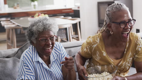 happy senior african american female friends eating popcorn, watching tv and laughing, slow motion