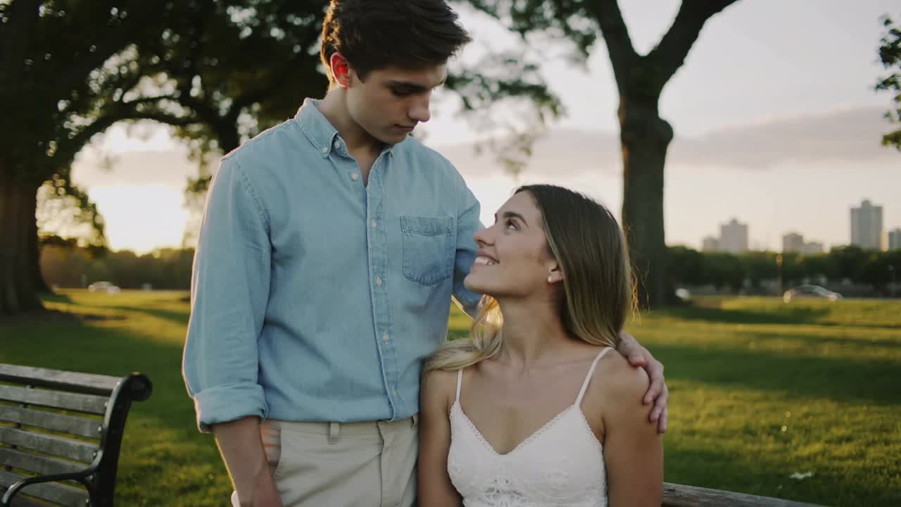 Homem com a mão sobre a mulher, observando-a e demonstrando amor, em um banco de parque ao pôr do sol, com ela usando um vestido branco.