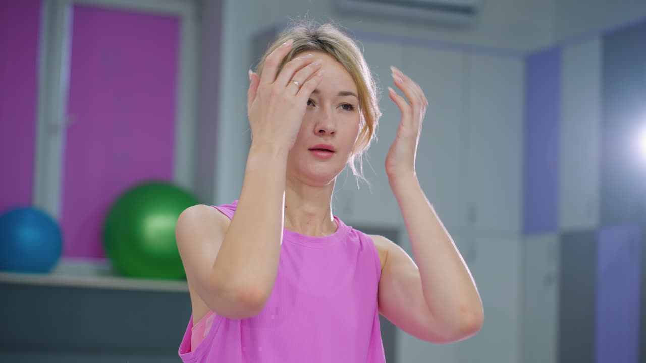 보라색 스포츠 옷을 입은 여성이 체육관에 서서 머리를 조정하고 배경에는 운동 공이 있습니다. 에서 빛나는 밝은 빛은 피트니스 스튜디오 분위기를 향상시니다.
