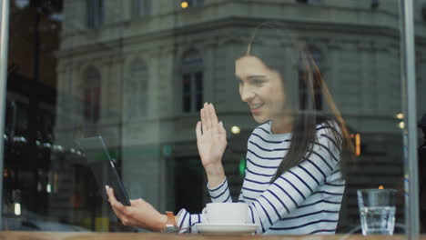 mujer joven morena sentada en el café y teniendo videochat en la tableta, hablando y siendo alegre