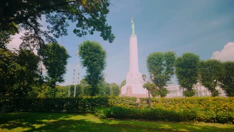 view from a park to the tall white freedom monument in riga, latvia, during a bright summer day, framed by lush greenery and trees