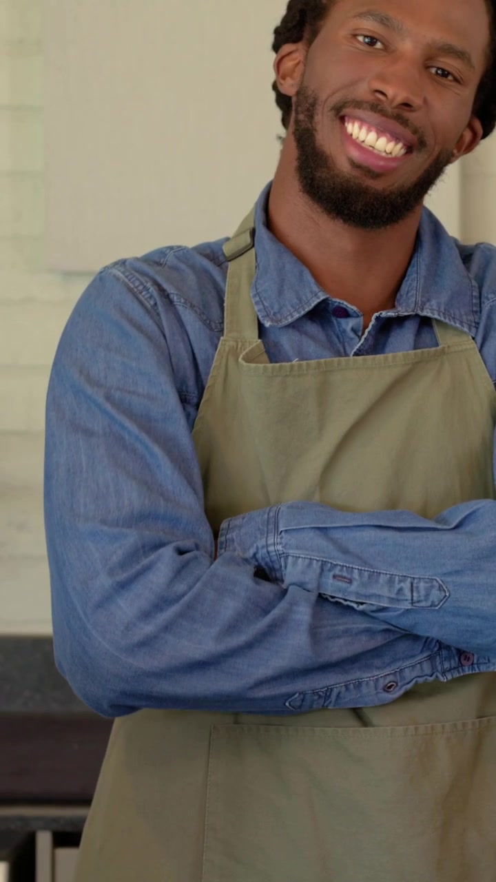 Vídeo vertical: Homem sorrindo com camisa jeans e avental, verificando um tablet no balcão de um café, enquanto atende um cliente.
