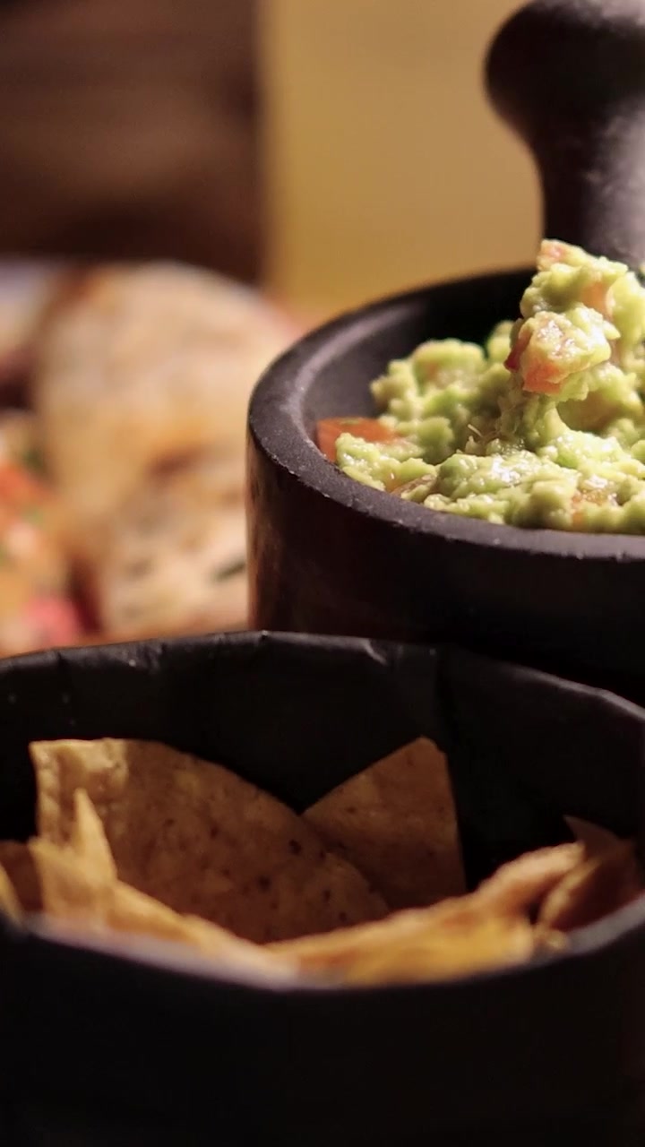 Una mano taglia una quesadilla grigliata su un piatto colorato, con guacamole e tortilla chips in ciotole rustiche sotto un'illuminazione calda e soffusa. La telecamera sposta lentamente il fuoco.