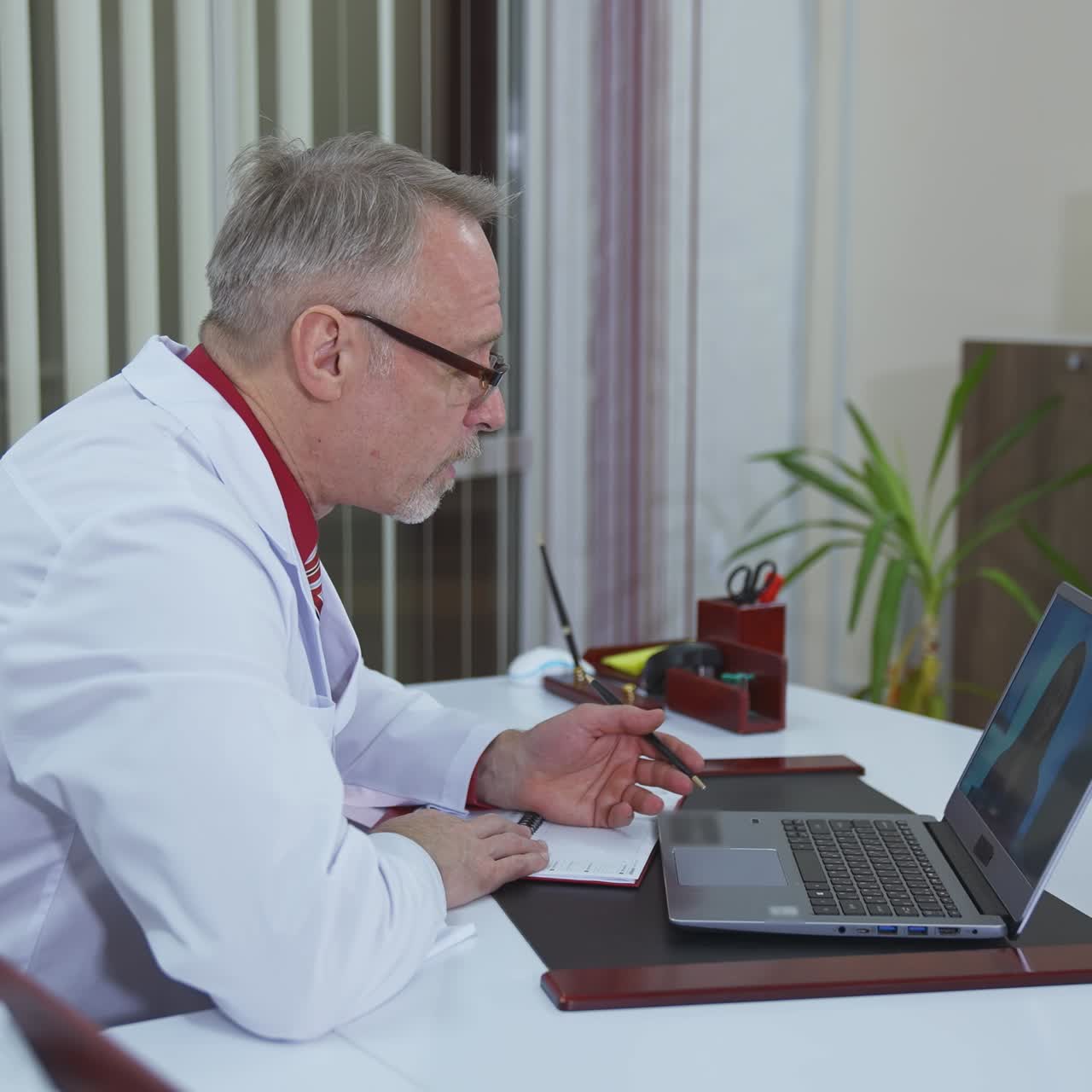 Doctor listens to a patient through the remote webcam on laptop screen. Professional male doctor in hospital office talking to a client online by video call on computer. Profile view.