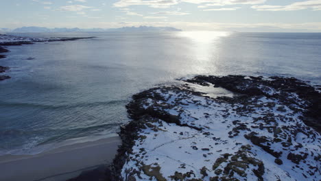 Truck-left-aerial-shot-of-a-snow-covered-rocky-outcrop-into-the-ocean-and-beach-in-Norway-with-bright-glare-off-the-ocean