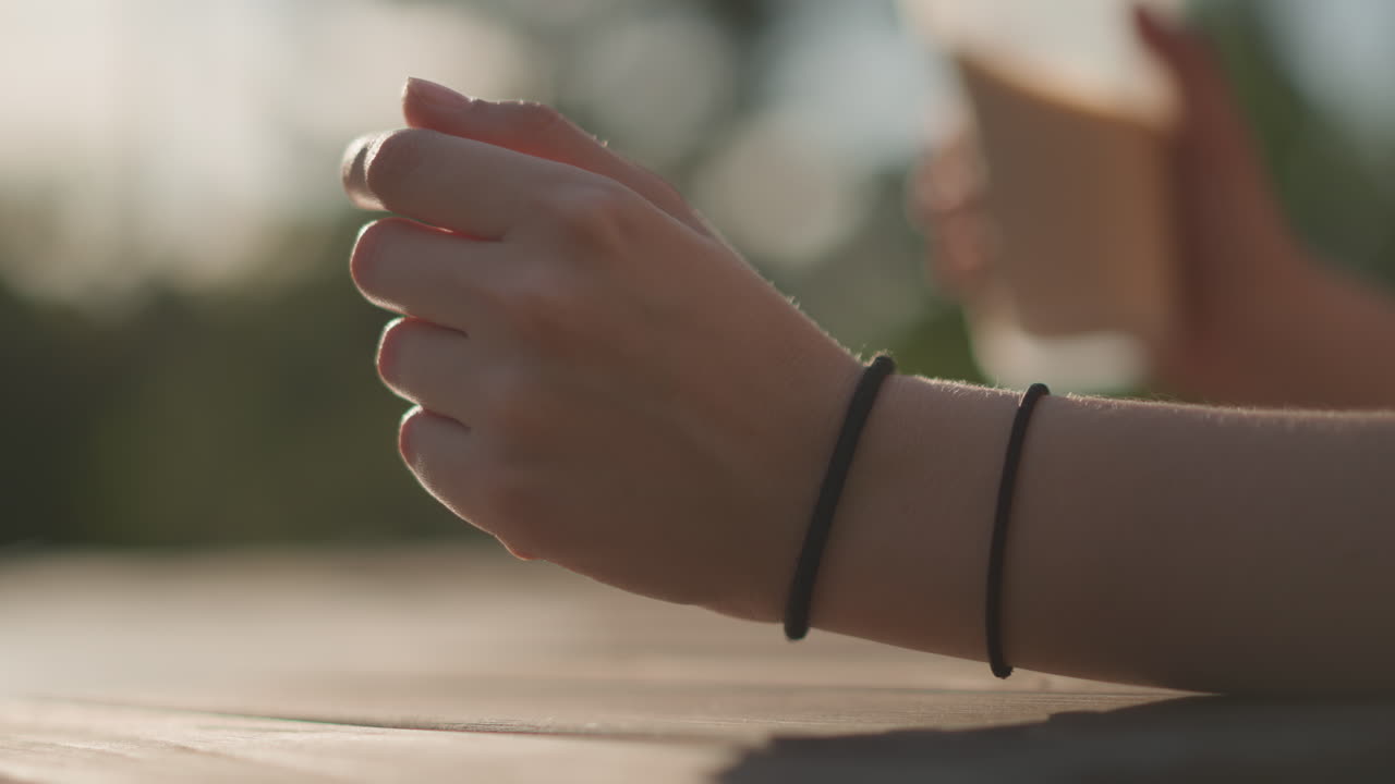 closeup wrist reaching for iced coffee cup, hand with elastic band lifts plastic cup, gentle grip and sip motion, sunlit picnic table with blurred park background, cinematic shallow focus and warm