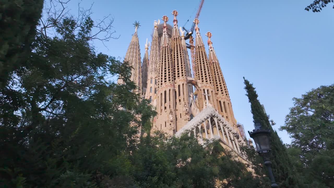 Green trees partially obscure Sagrada Familia in Barcelona, Spain, with construction crane visible against blue sky