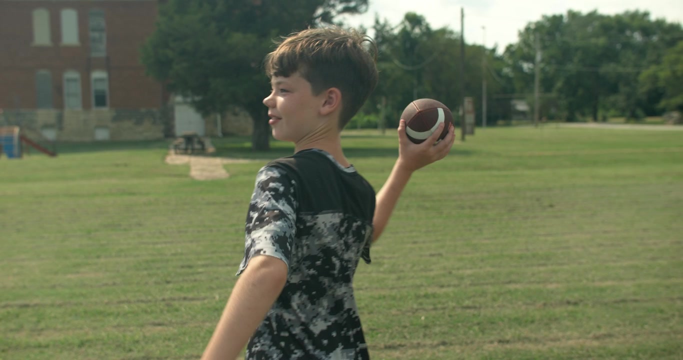 Young boy holds and throws an American football while playing outdoors on an open grassy field.