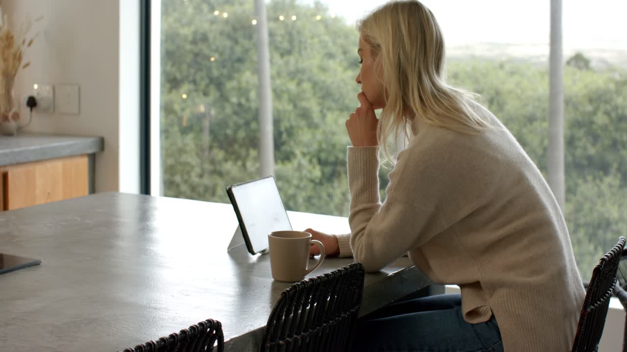 After propping tablet upright, woman tapping screen, leaning by window and bringing mug closer