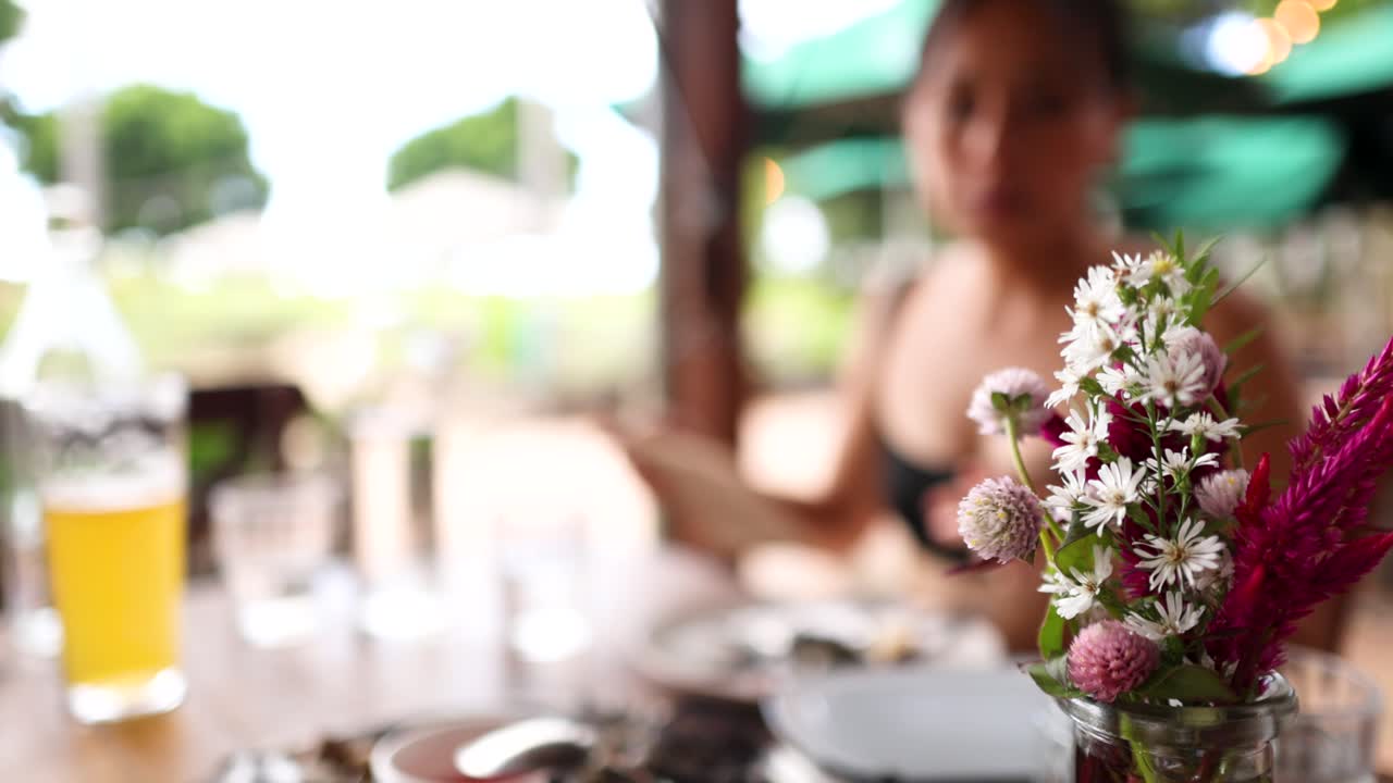 A dining table with flowers in focus, blurred person in background, bright outdoor lighting