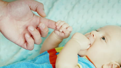 bebé despreocupado de dos meses mordiéndose el puño sosteniendo el dedo de la mano de su padre acostado en la cama feliz