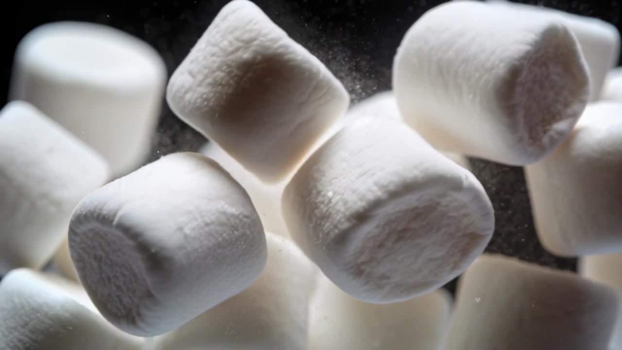 Marshmallow che cadono, ruotano e si separano in studio dopo un soffio, mostrando polvere sospesa.