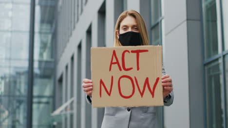 portrait of caucasian elegant young woman in facial mask showing act now" signboard in the street"