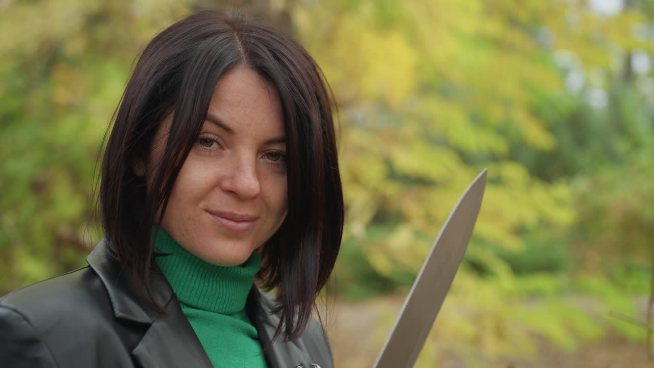 Una mujer con una chaqueta de cuero inspecciona un cuchillo en un claro de un bosque otoñal, con un jersey de cuello alto verde que contrasta con el follaje amarillo, con un termo y un tronco cerca. Examina la hoja con una expresión tranquila y confiada, primer plano.