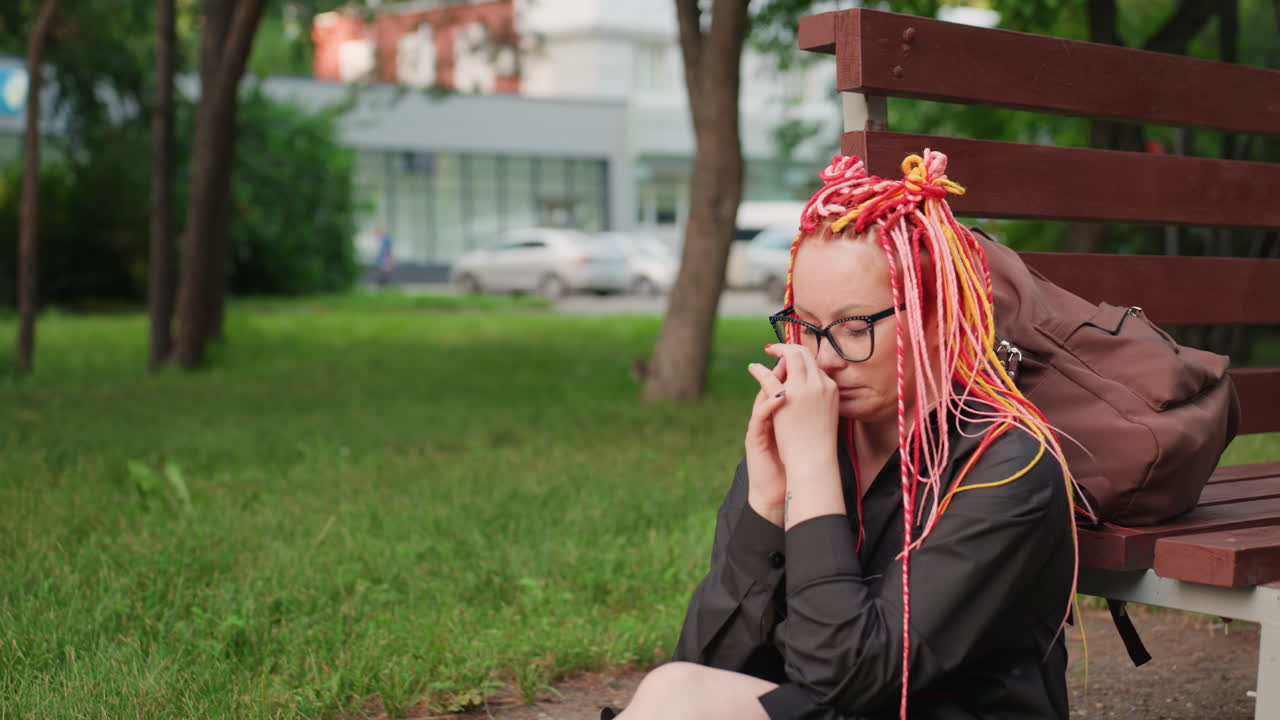 분홍색 머리를 한 젊은 여성, 분홍색 드레드락을 한 여성이 도시 공원에서 걱정하는 듯한 모습으로, 분홍색 드레드락을 한 여성이 도시 환경과 분주한 교통 속에서 녹색 잔디에 앉아 있어요.