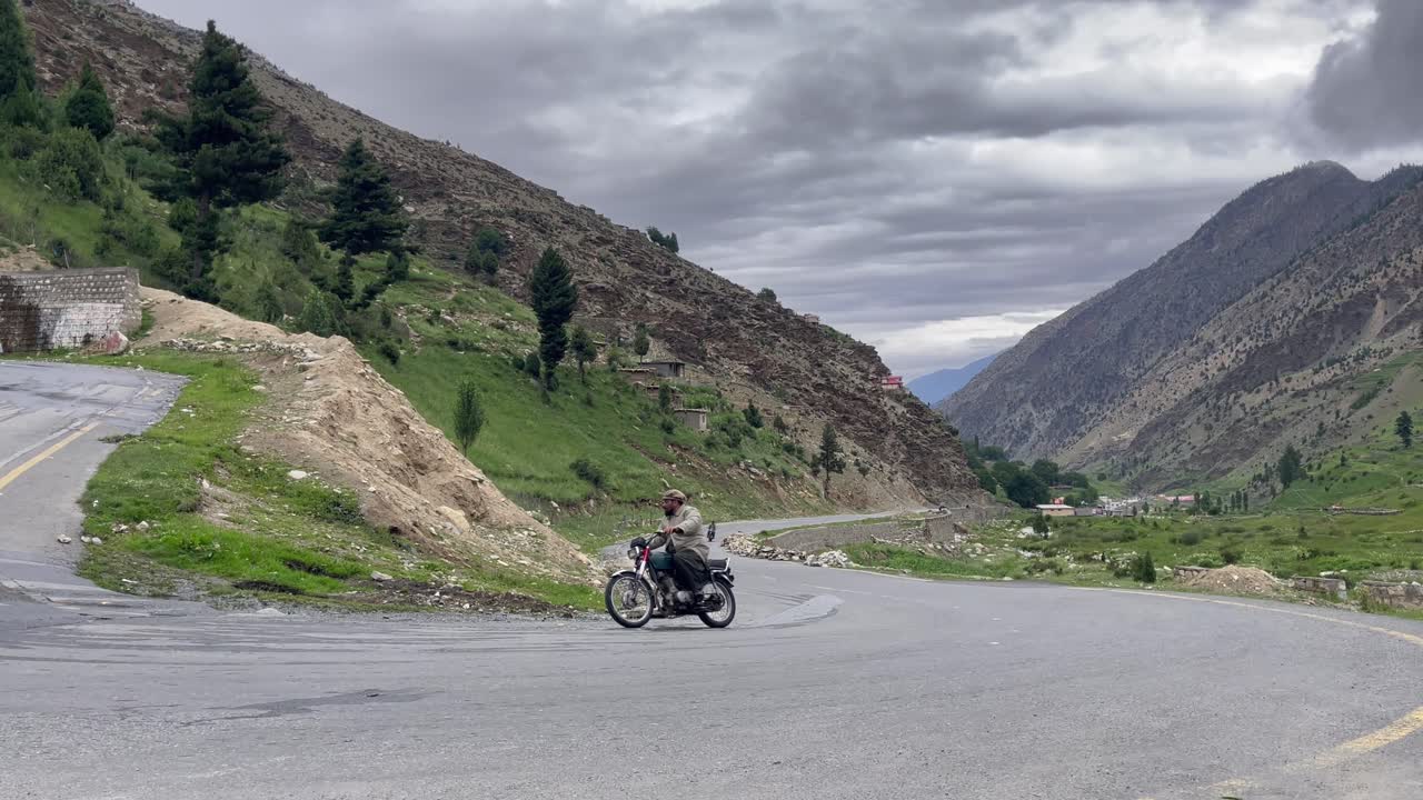 A motorbike rider confidently takes on the scenic and challenging winding mountain road at Babusar Pass, connecting Khyber Pakhtunkhwa with Gilgit-Baltistan under a dramatic, cloudy sky.