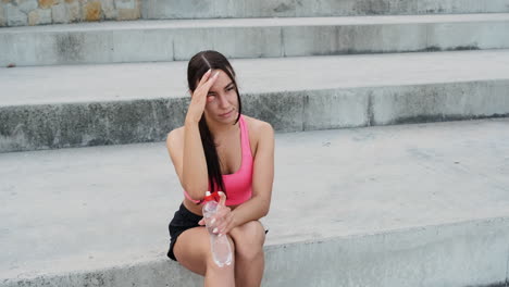 bella deportista sentada en las escaleras y bebiendo agua después de una sesión de entrenamiento en la ciudad