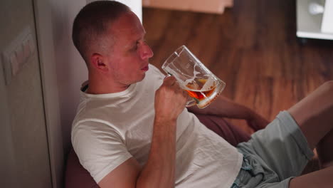 man sitting casually on floor against wall drinking beer from transparent glass mug wearing white t shirt and light shorts in cozy indoor setting with wooden floor