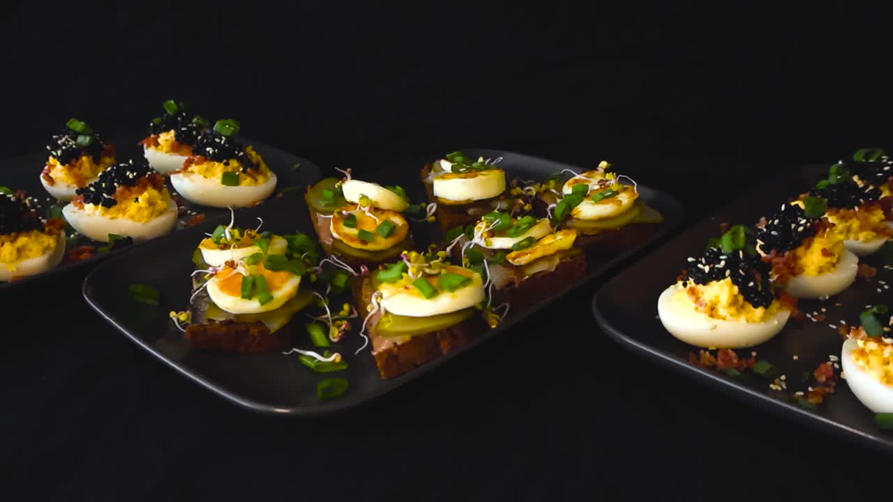 Photo en studio d'amuse-gueules estoniens traditionnels, notamment des œufs mimosa garnis de caviar et de bacon, et des canapés de pain de seigle avec des tranches d'œufs de sprat, des cornichons et des micropousses, sur fond sombre.