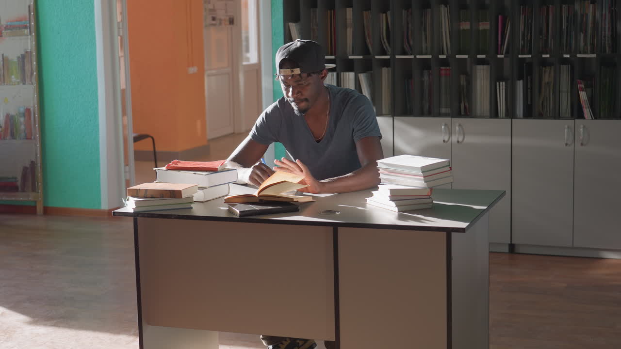 밝은 도서관 책상에 앉아 모자를 쓴 남자가 책을 펼쳐 새로운 페이지를 보며 필기를 하고 있고, 주변에는 여러 겹의 책들이 쌓여 있으며, 큰 창문으로는 햇빛이 쏟아져 들어오고 있어요.