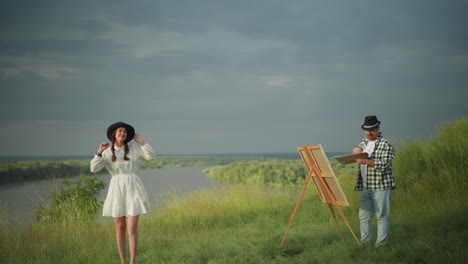 an artist holding a palette and brush paints a woman in a white dress and hat who stands poised in front of him. the scene unfolds under a cloudy sky, with a serene lakeside