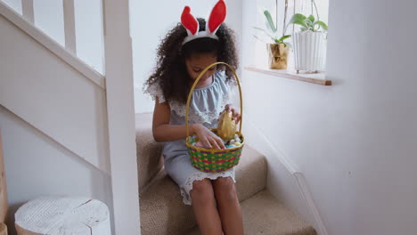 Girl-Wearing-Bunny-Ears-Sitting-On-Stairs-Eating-Chocolate-Egg-She-Has-Found-On-Easter-Egg-Hunt