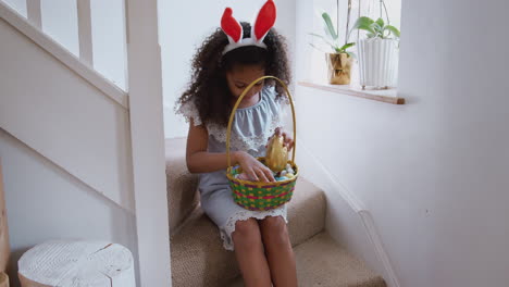 girl wearing bunny ears sitting on stairs eating chocolate egg she has found on easter egg hunt