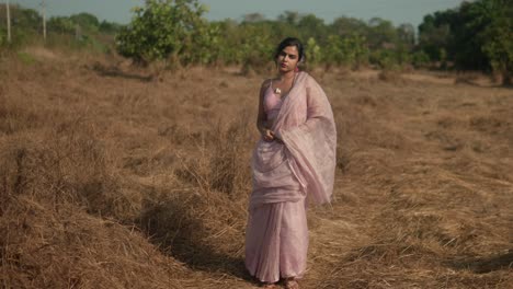 woman in traditional sari standing contemplatively in dry grassland, with trees in the background, daylight