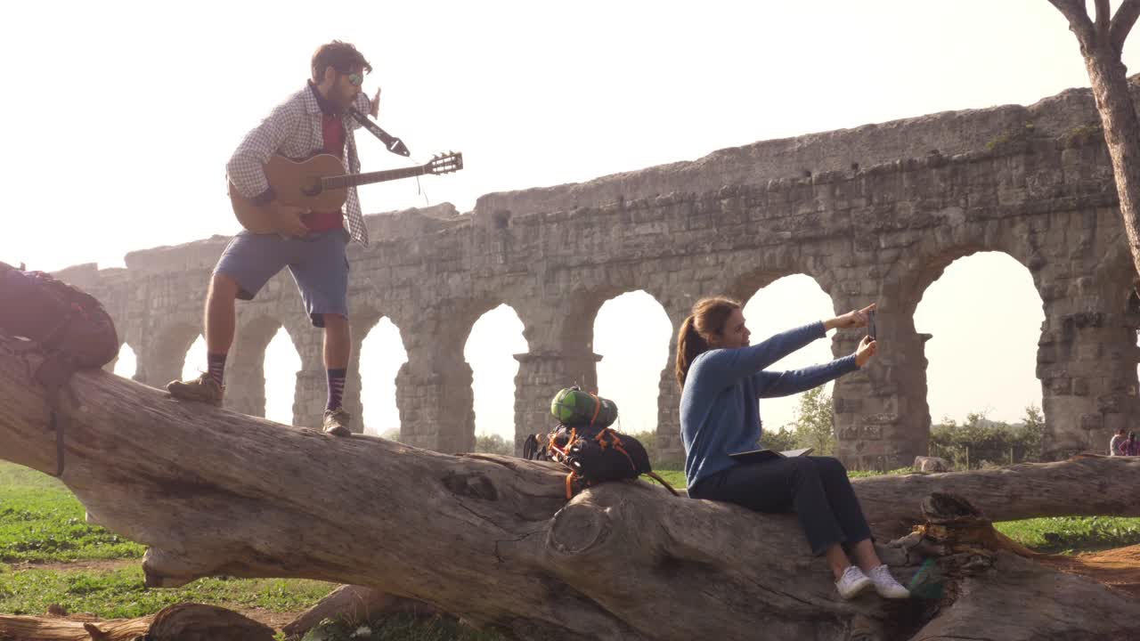 행복한 젊은 부부, 배 여행자, 트크에 앉아 스마트폰으로 셀카를 찍는 관광객, 로마의 만적 인 파르코 델리 아쿠에도티 공원의 고대 로마 수로 잔해 앞에서, 안개가 은 해가 뜨는 느린 동작.