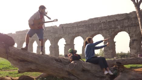 happy young couple backpackers tourists on a log trunk taking selfies photos with smartphone in front of ancient roman aqueduct ruins in romantic parco degli acquedotti park in rome misty sunrise slow motion