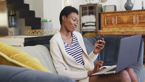 Feliz-Mujer-Afroamericana-Sentada-En-Un-Sofá-En-La-Sala-De-Estar,-Usando-Una-Computadora-Portátil-Y-Un-Teléfono-Inteligente